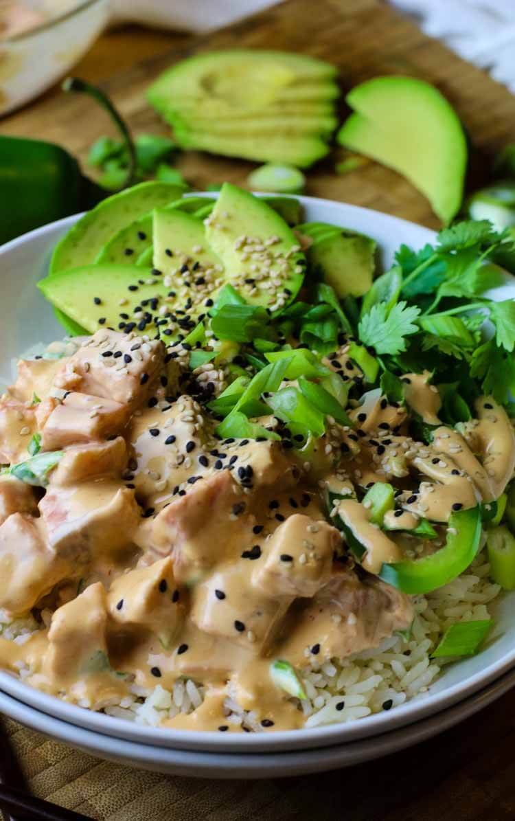 Salmon poke in a white bowl on a bed of white rice with avocado, cilantro and green onions topped.