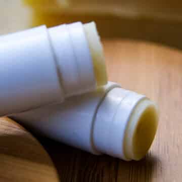 Homemade lip balm (2 tubes) that are opened on a bamboo cutting board with beeswax bricks in the background.