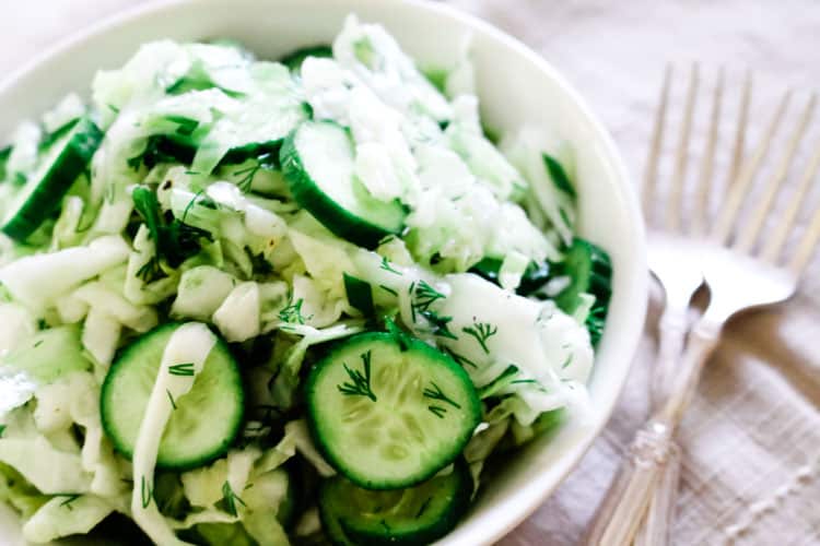 white bowl with sliced cucumbers, shredded cabbage and two forks off to the side