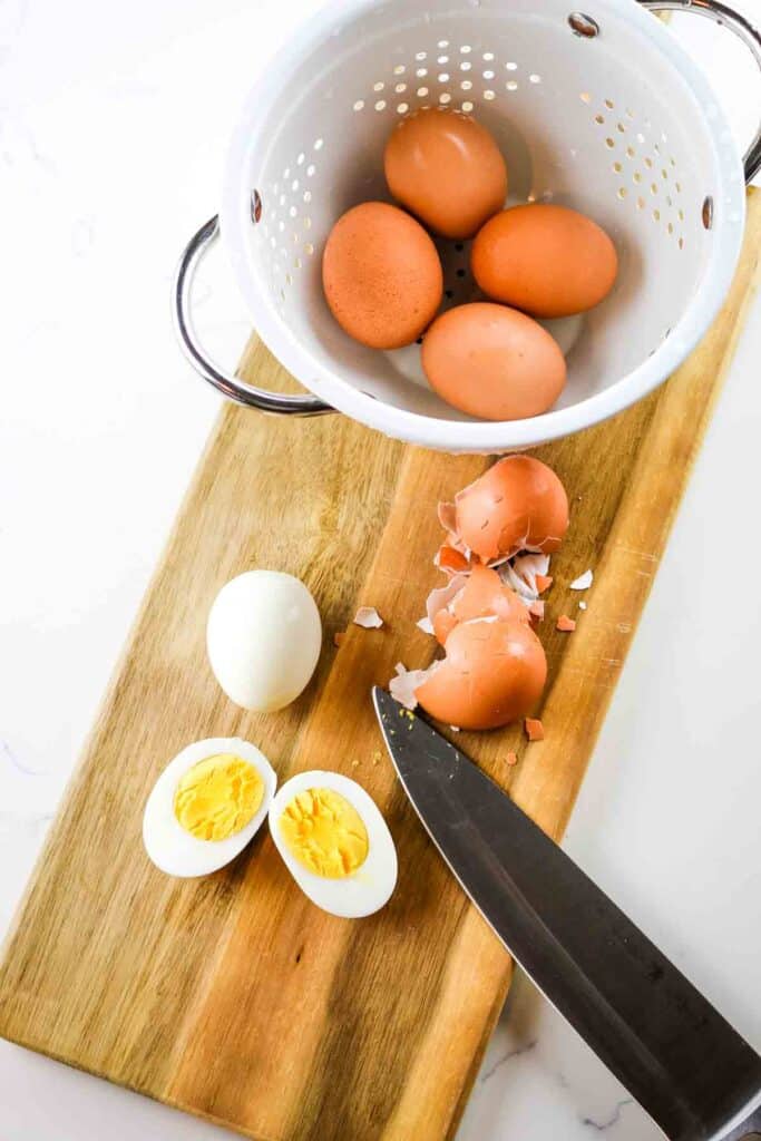 Hard boiled eggs on a cutting board with knife and colander, with one egg peeled and cut in half.