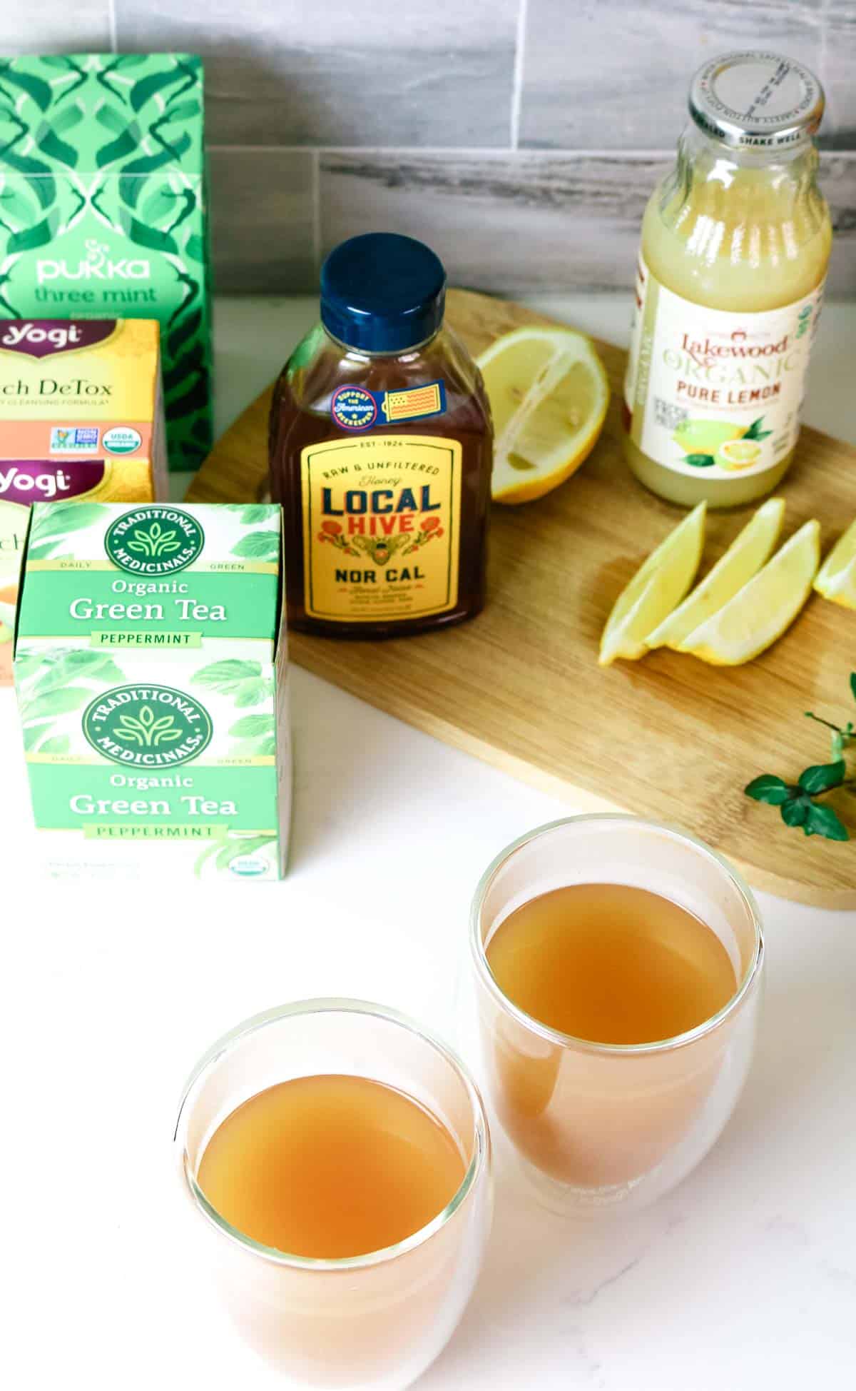 Image of 2 mugs of Honey Citrus Mint Tea and tea boxes, honey, lemon juice and wedges in the background