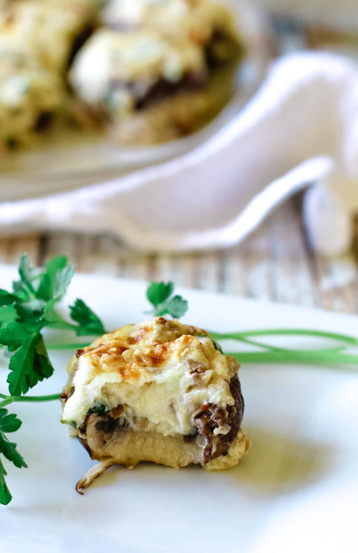 A stuffed mushroom on a white plate with platter of mushrooms in the background.