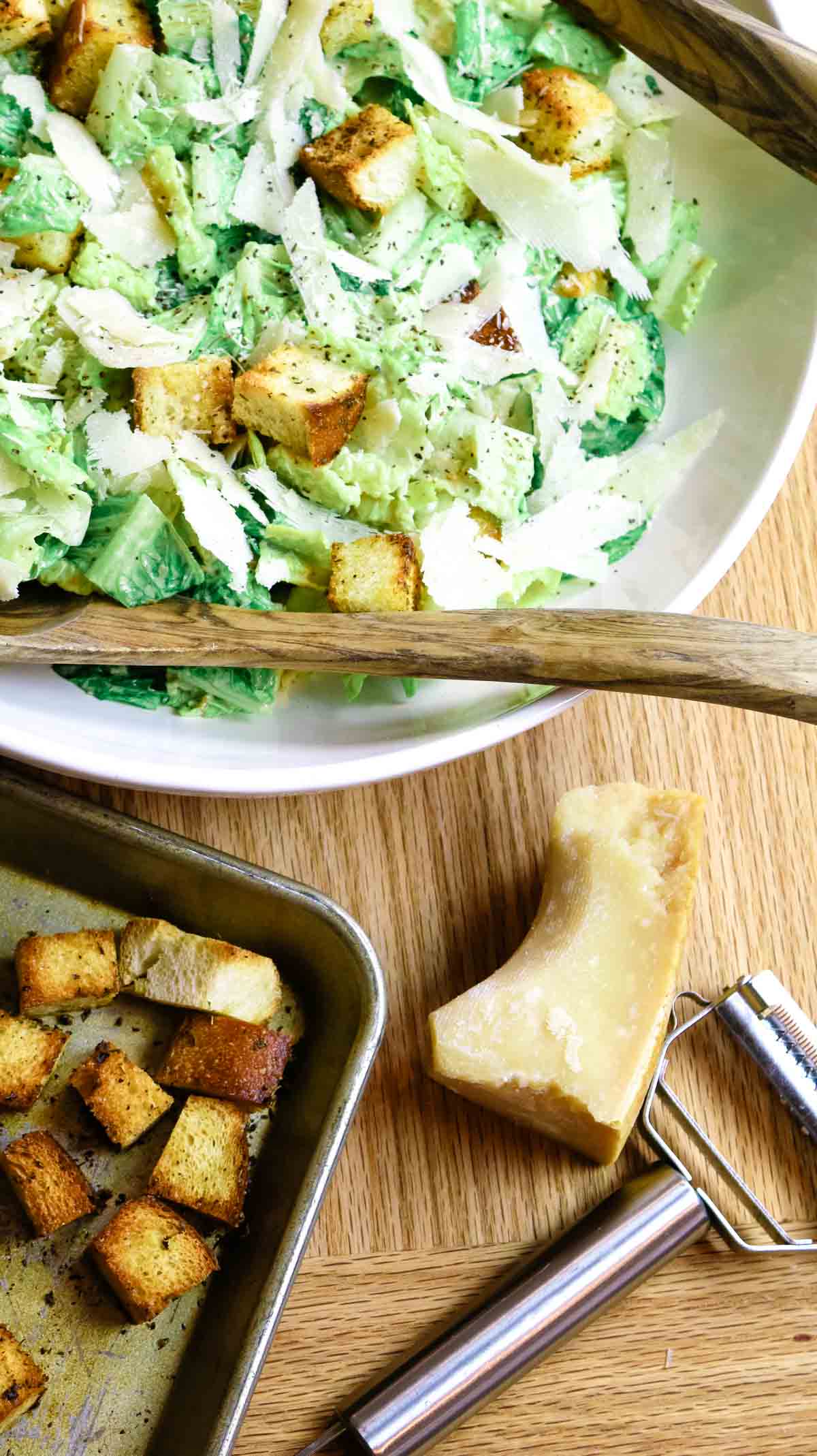 bowl with homemade caesar salad, baking sheet with croutons and fresh parmesan cheese with shaver