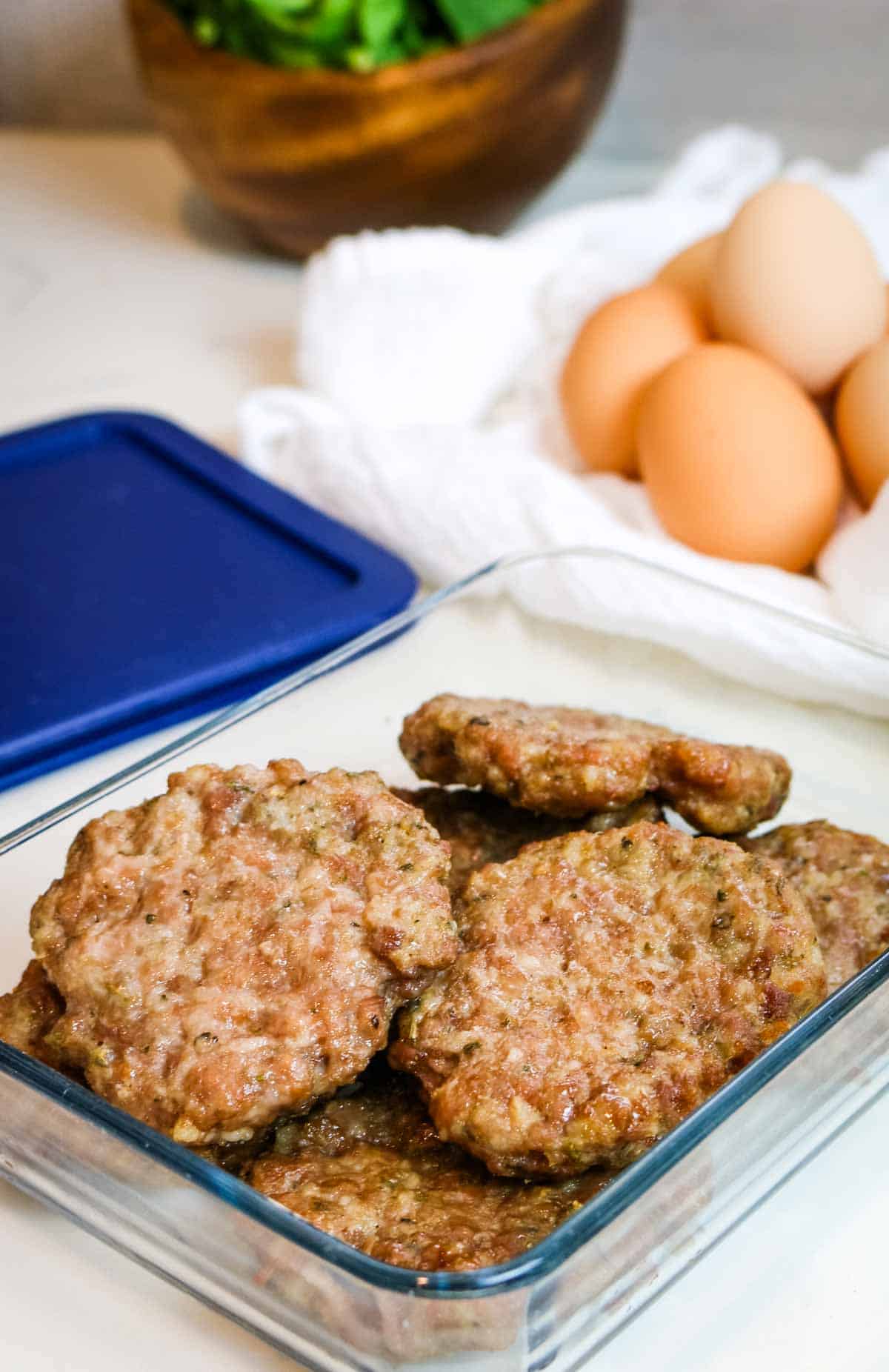 sausage patties in a glass storage container with eggs in the background
