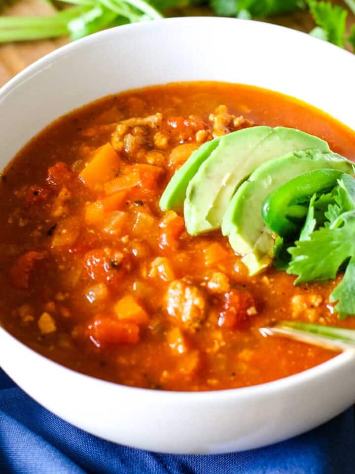 turkey chili with avocado and cilantro in white bowl and with blue napkin on side.