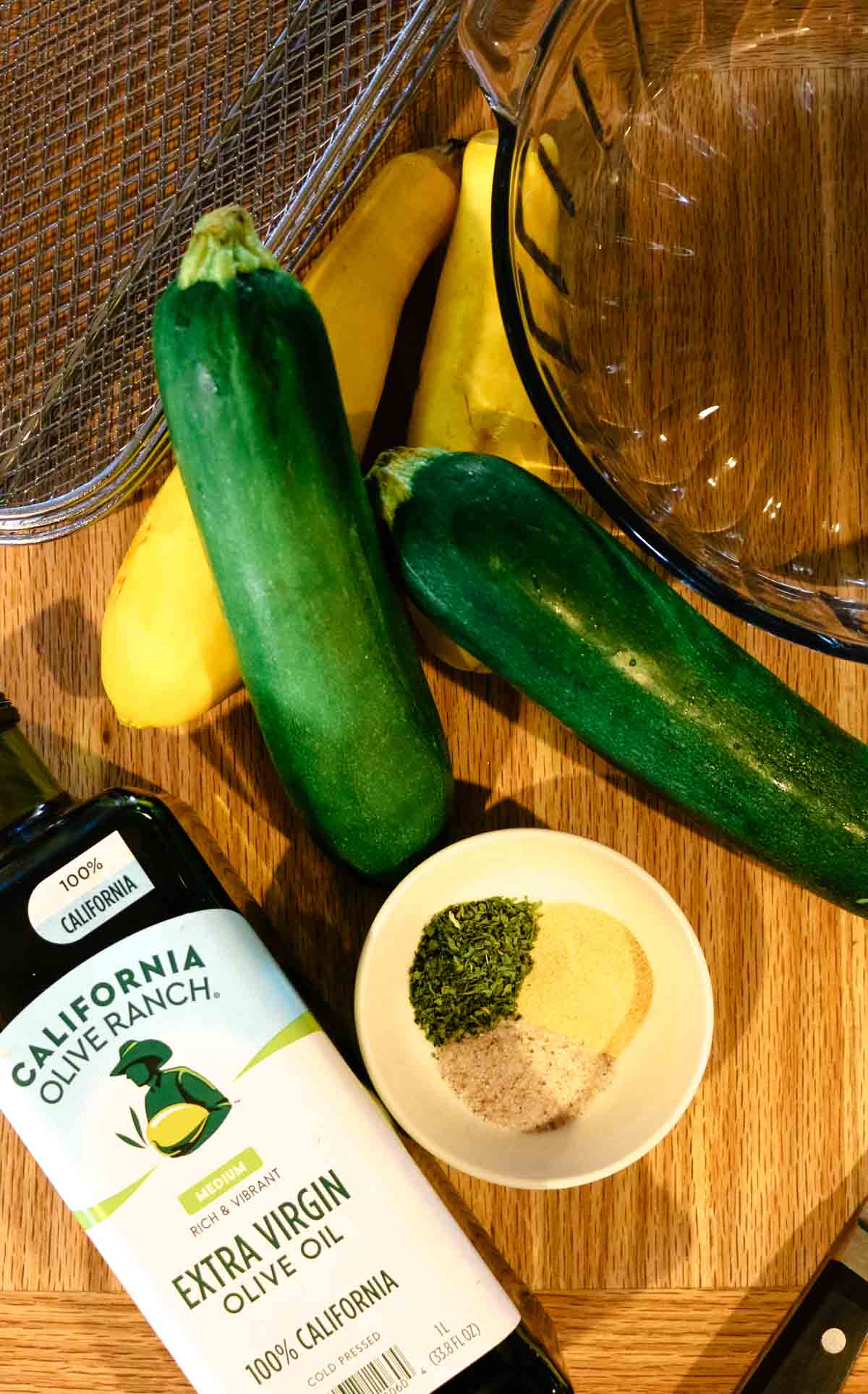olive oil, zucchini, and yellow squash with seasonings on a cutting board.