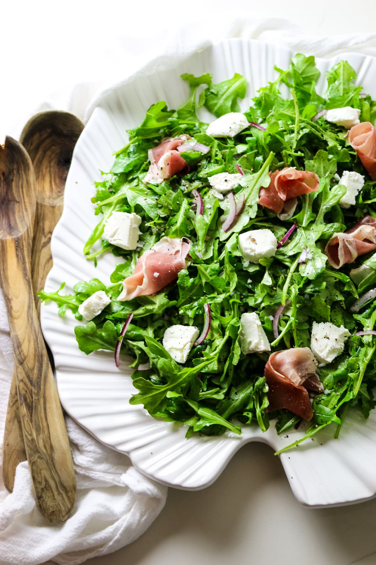 rocket salad with arugula leaves on a white platter with goat cheese and prosciutto toppings with wooden spoons on the left.