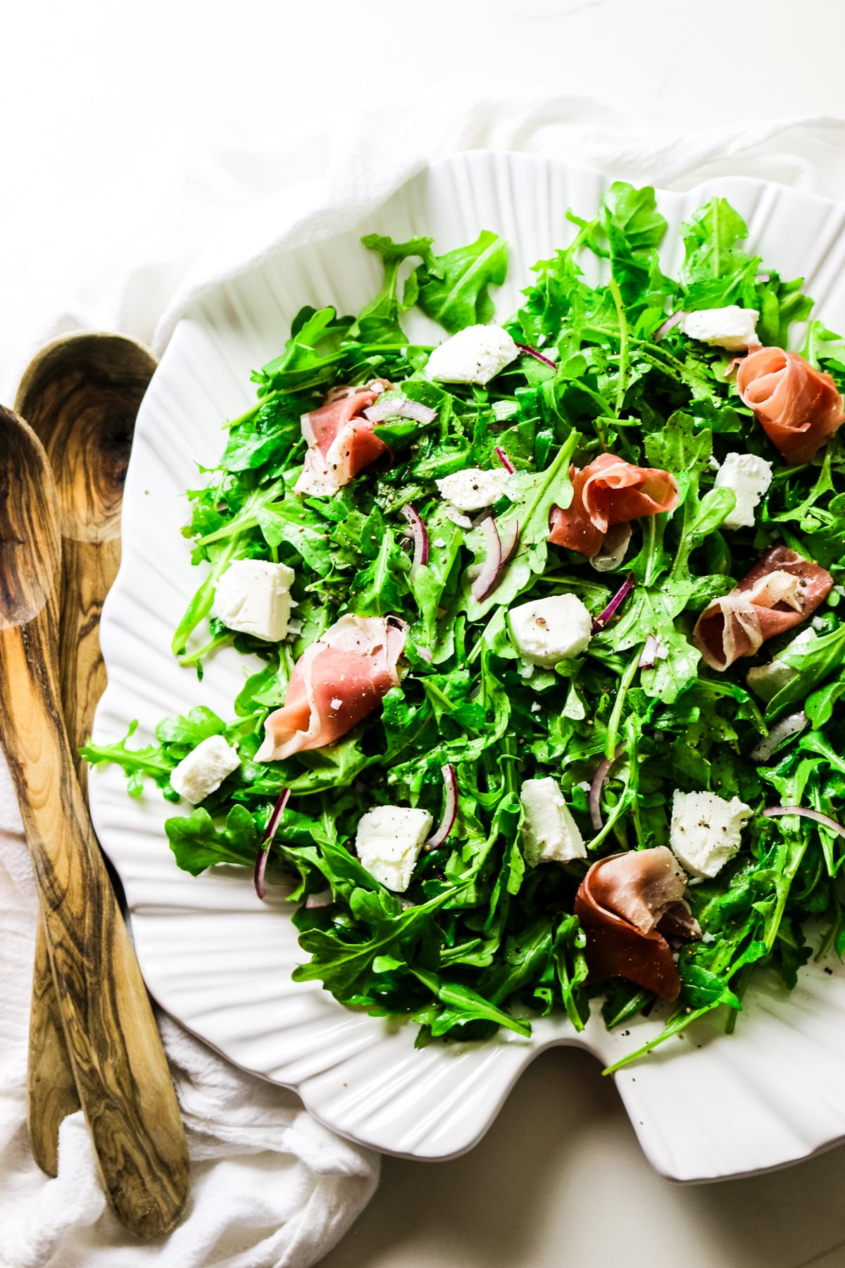 rocket salad with arugula leaves on a white platter with goat cheese and prosciutto toppings with wooden spoons on the left.