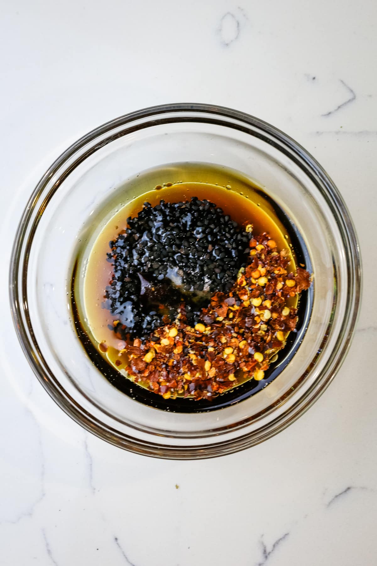 overhead of clear small bowl with dressing, black sesame seeds and red chili flakes.