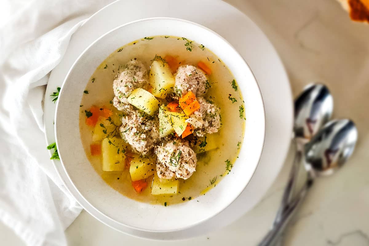 chicken meatball soup with potatoes and carrots in white bowl and white towel on the side.