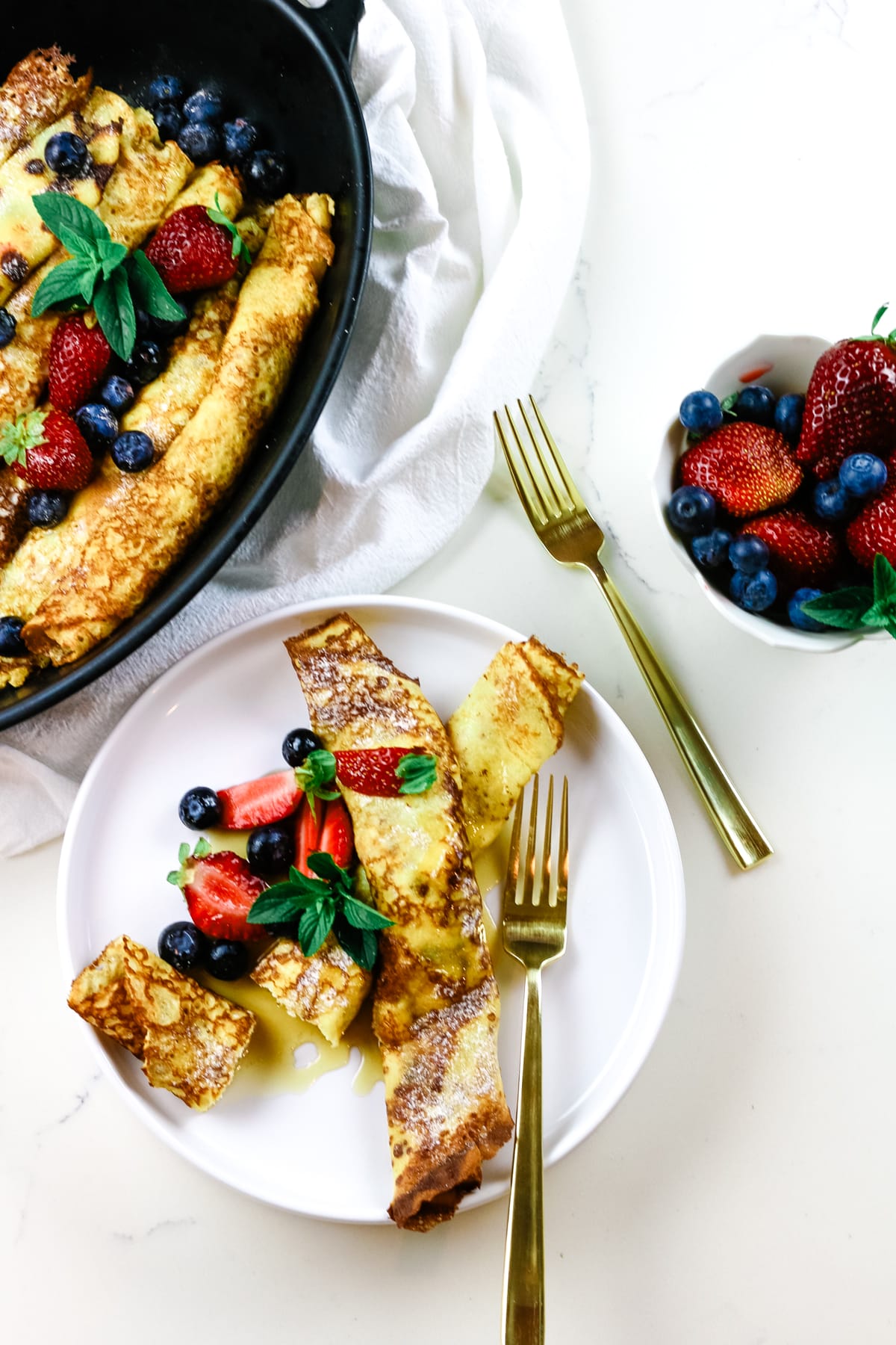 cottage cheese crepes on a white plate with baking dish with them on the side and fresh berries to the right.