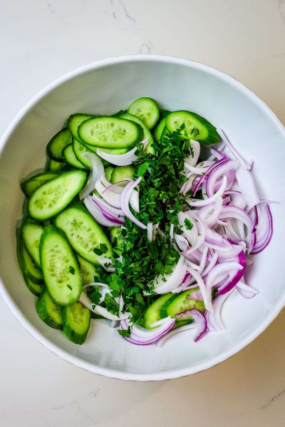 fresh ingredients added to white bowl including cucumbers, cilantro, and red onion.