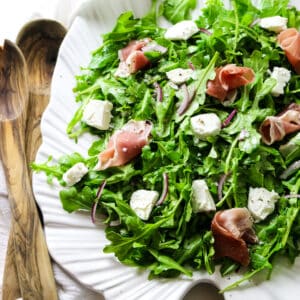 rocket salad with arugula leaves on a white platter with goat cheese and prosciutto toppings with wooden spoons on the left.