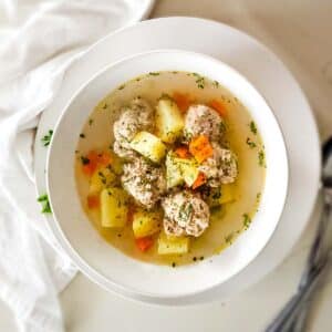meatball soup with potatoes and carrots in a very clear broth in white bowl.