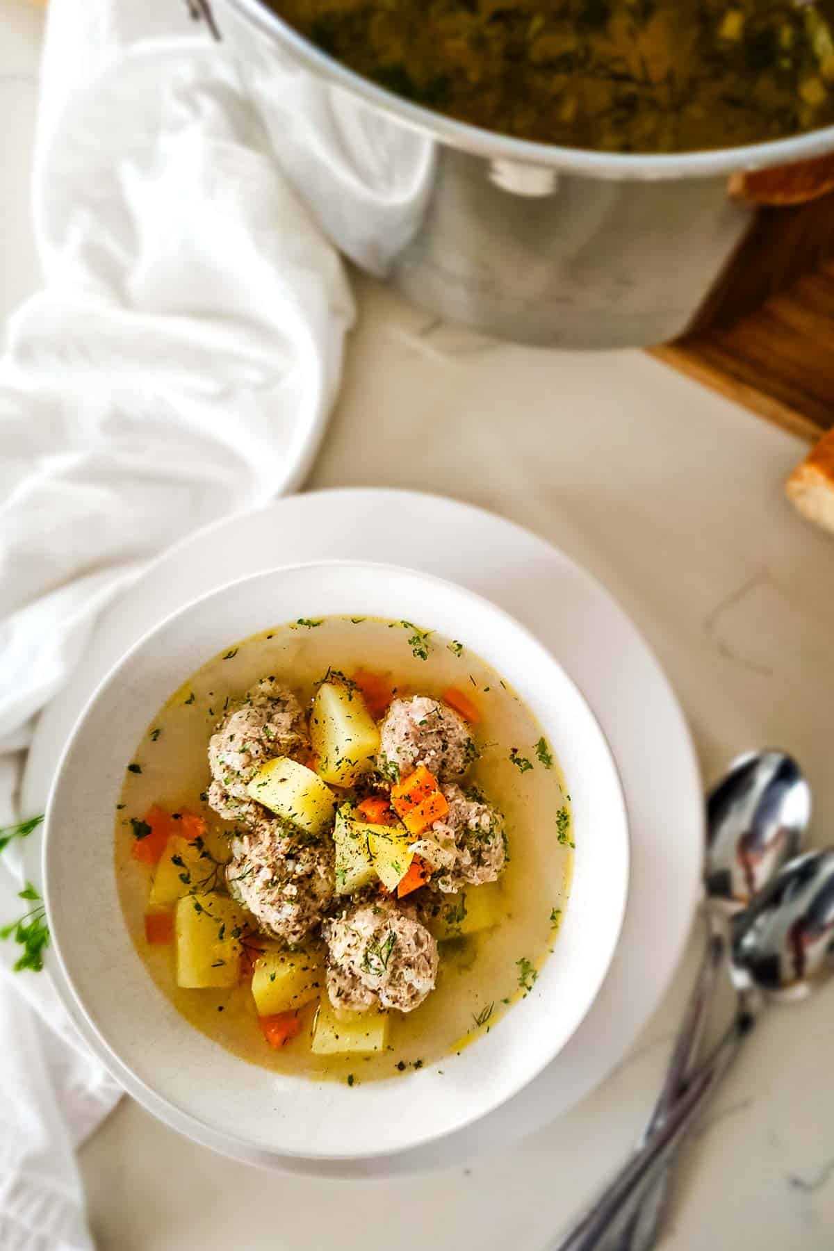 meatball soup in broth in white bowl and spoons to the right and large soup pot in the upper right corner.