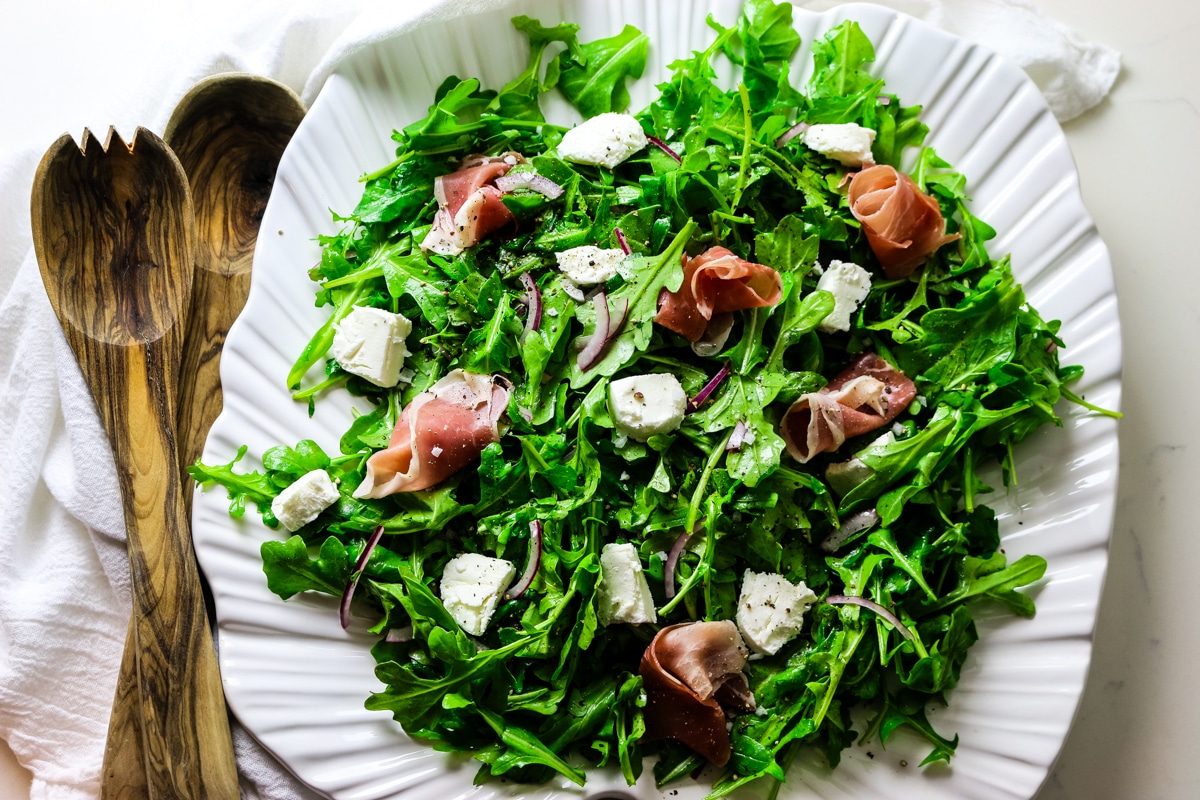 rocket salad with arugula leaves on a white platter with goat cheese and prosciutto toppings with wooden spoons on the left.