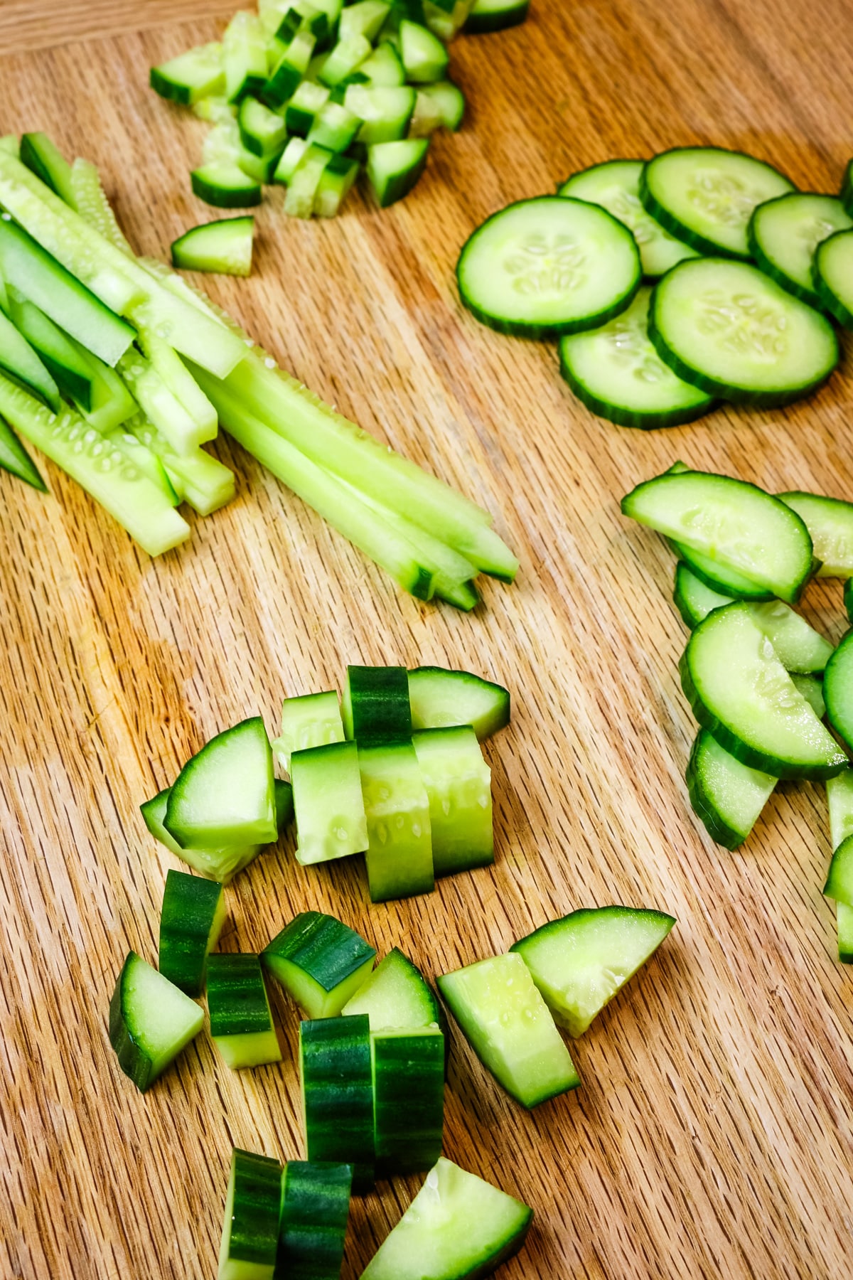 cucumber cut in many different shapes and sizes on cutting board.