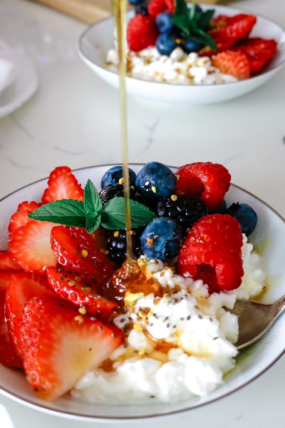 cottage cheese in a small bowl with sliced strawberries, raspberries, blueberries, blackberries, sprig of mint and honey being drizzled over the top.