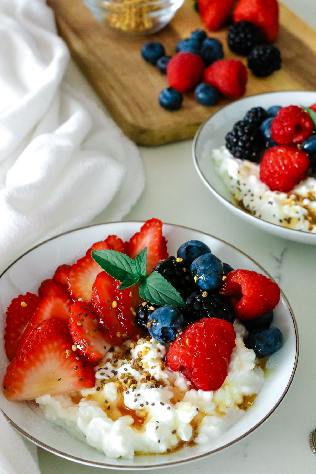 cottage cheese in a bowl with fresh mixed berries, sprig of mint, bee pollen and drizzled with honey and white kitchen towel to the left.