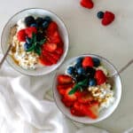 two bowls filled with cottage cheese and fresh strawberries, raspberries, blueberries, and blackberries and topped with honey; spoons in the bowls.