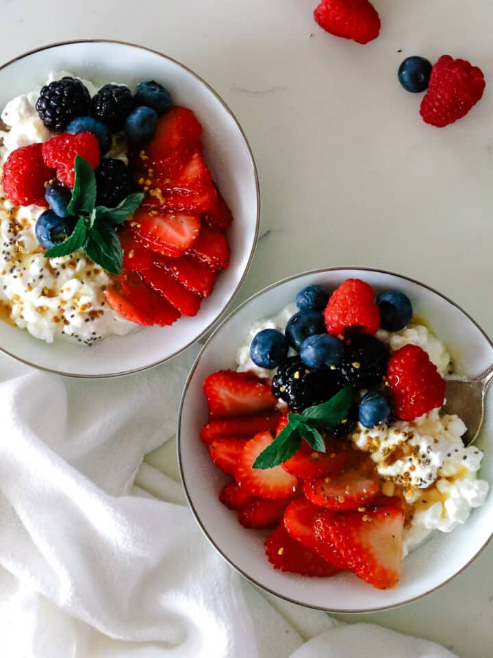 two bowls filled with cottage cheese and fresh strawberries, raspberries, blueberries, and blackberries and topped with honey; spoons in the bowls.