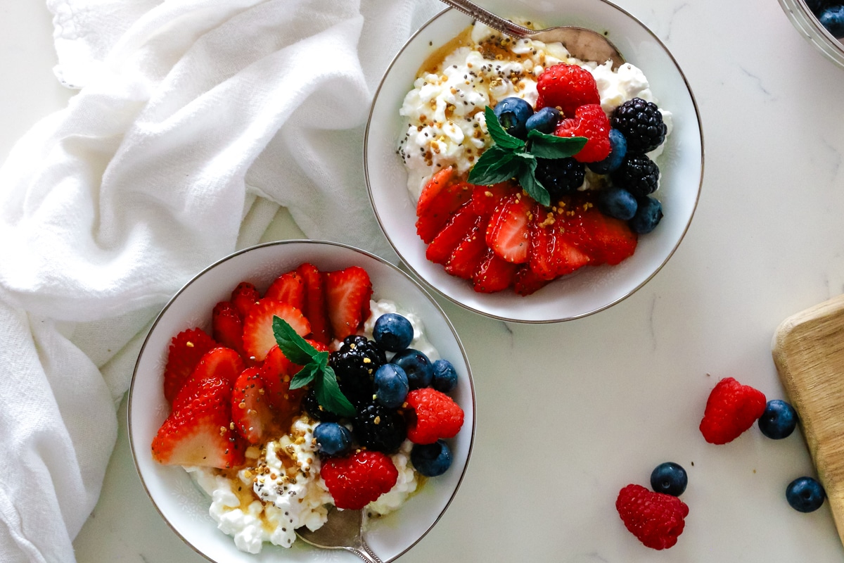 cottage cheese in bowls with fresh sliced berries and honey drizzle, chia seeds, and bee pollen on top and spoons inserted into the bowls.
