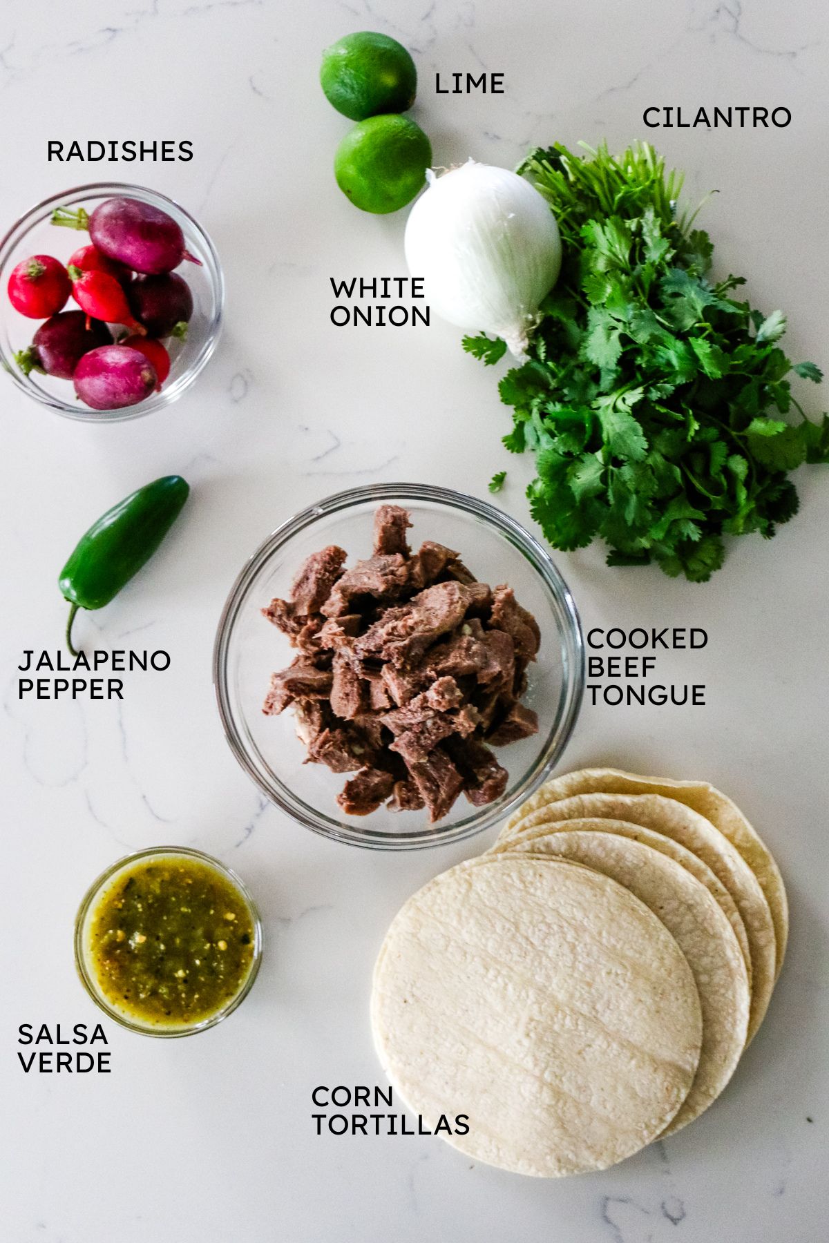 Ingredients on a white counter to make beef tacos, including cilantro, lime, radishes, pepper, beef tongue, salsa and corn.