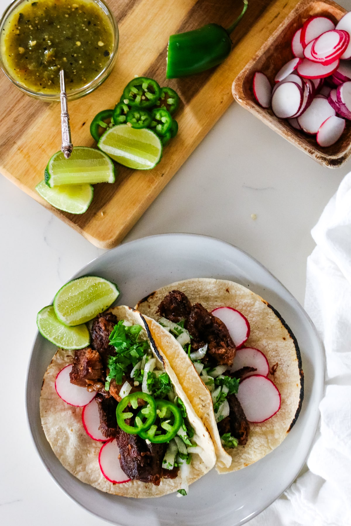 Two cow tongue tacos on white platter next to lime and cutting board with ingredients.