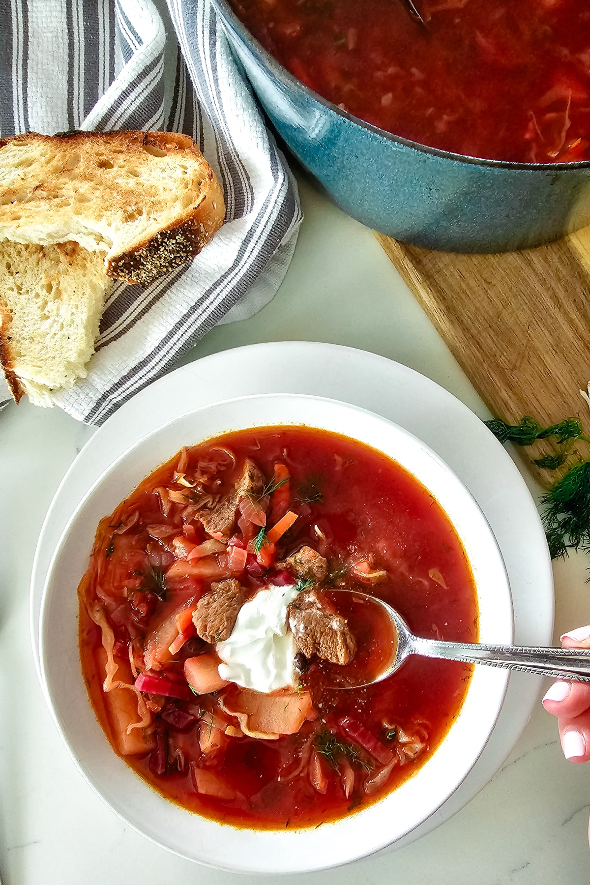 borscht in a white bowl on a white plate with bread to the left of it.
