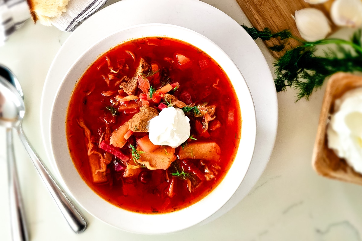 russian beet soup in a white bowl over white plate with a dollop of sour cream.