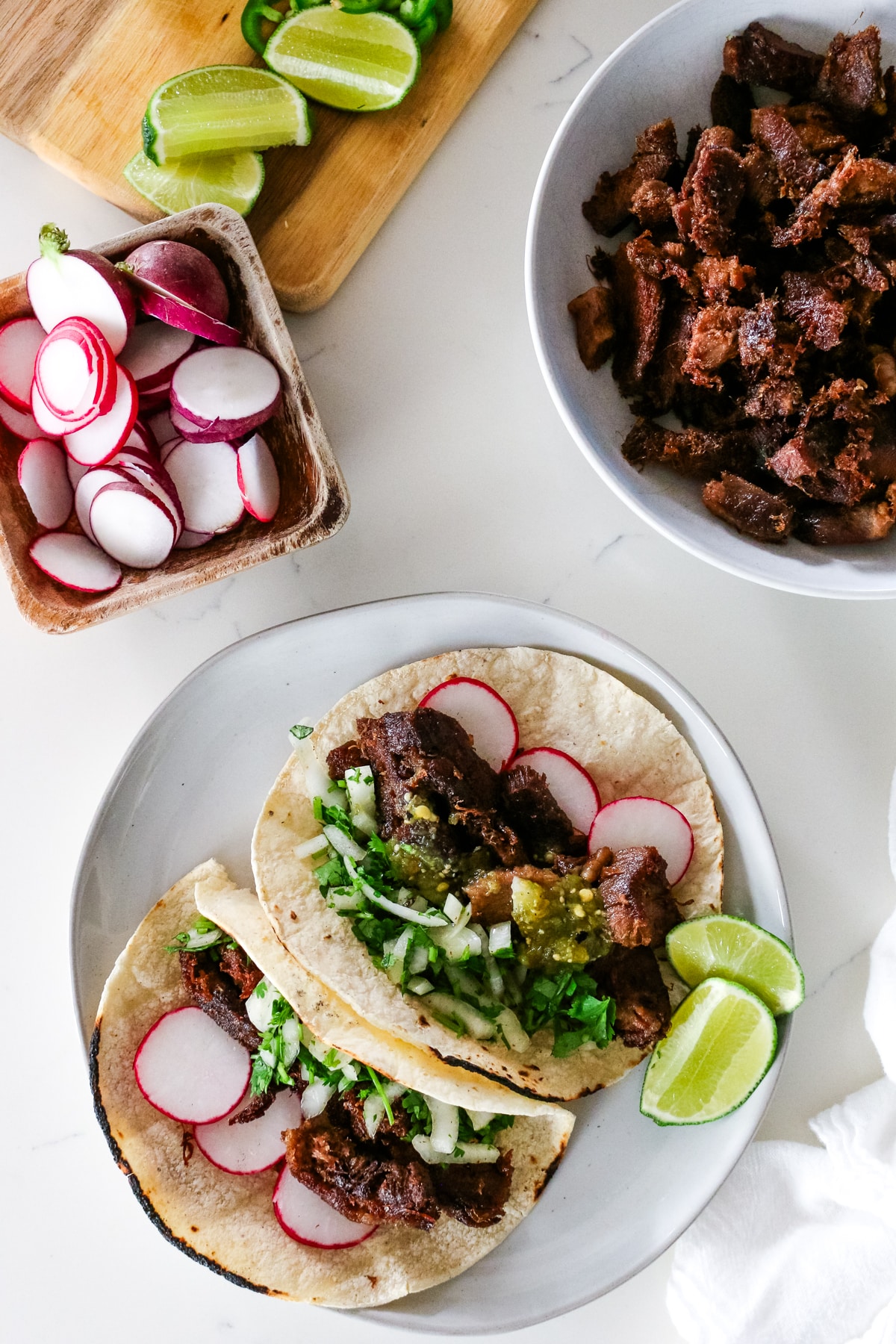 Two cow tongue tacos on white platter next to lime and cutting board with ingredients.