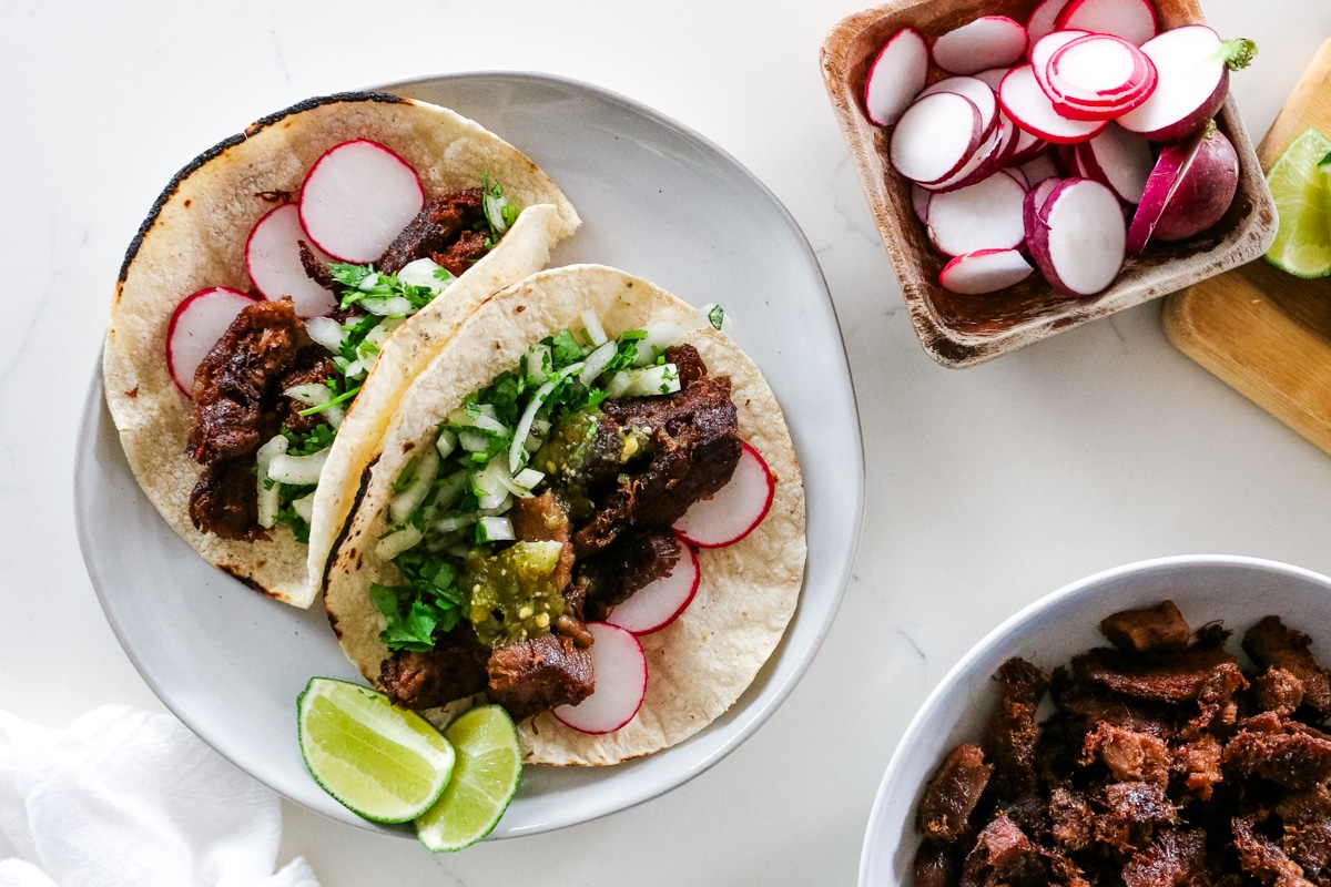 two beef tongue tacos on platter with radish in a wooden bowl and extra beef tongue in a platter below.