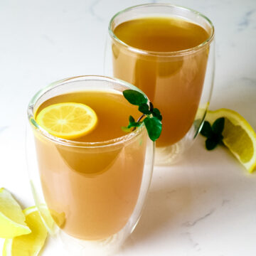 two clear mugs with honey citrus mint tea and lemon slices, lemon wedges, and fresh mint as garnish.