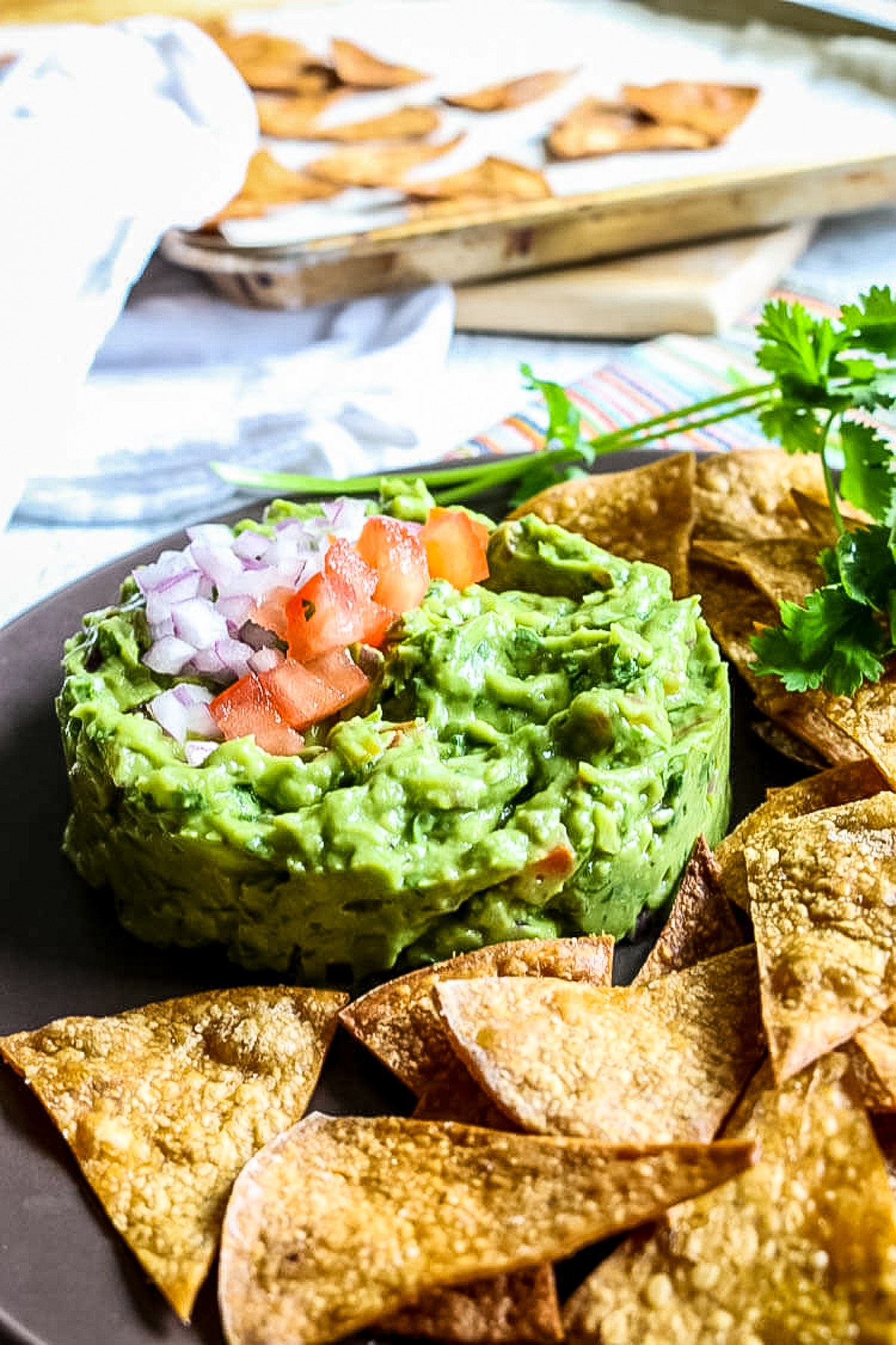 tortilla chips laying on a brow platter next to guacamole, in the back a baking sheet has freshly made chips.