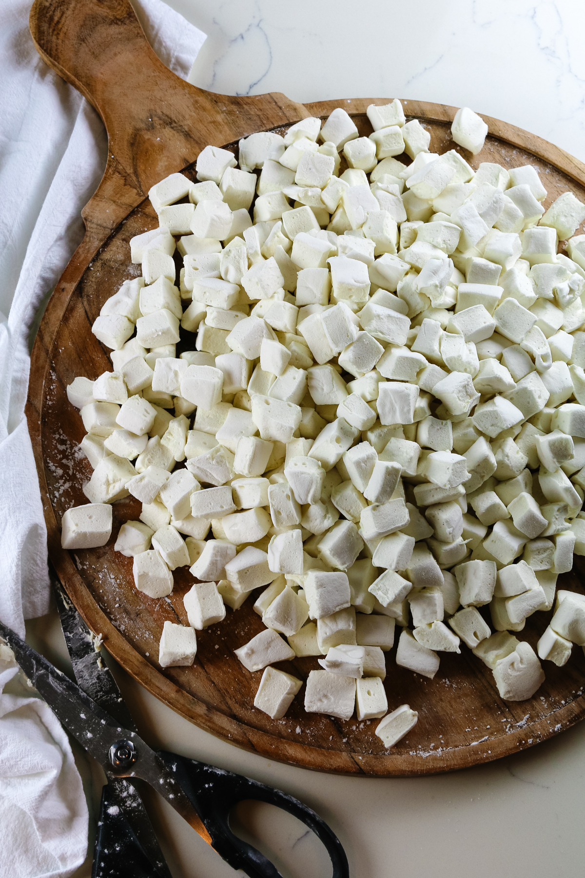 homemade marshmallows cut into bite-size pieces on a circular wooden cutting board and kitchen shears to the side.
