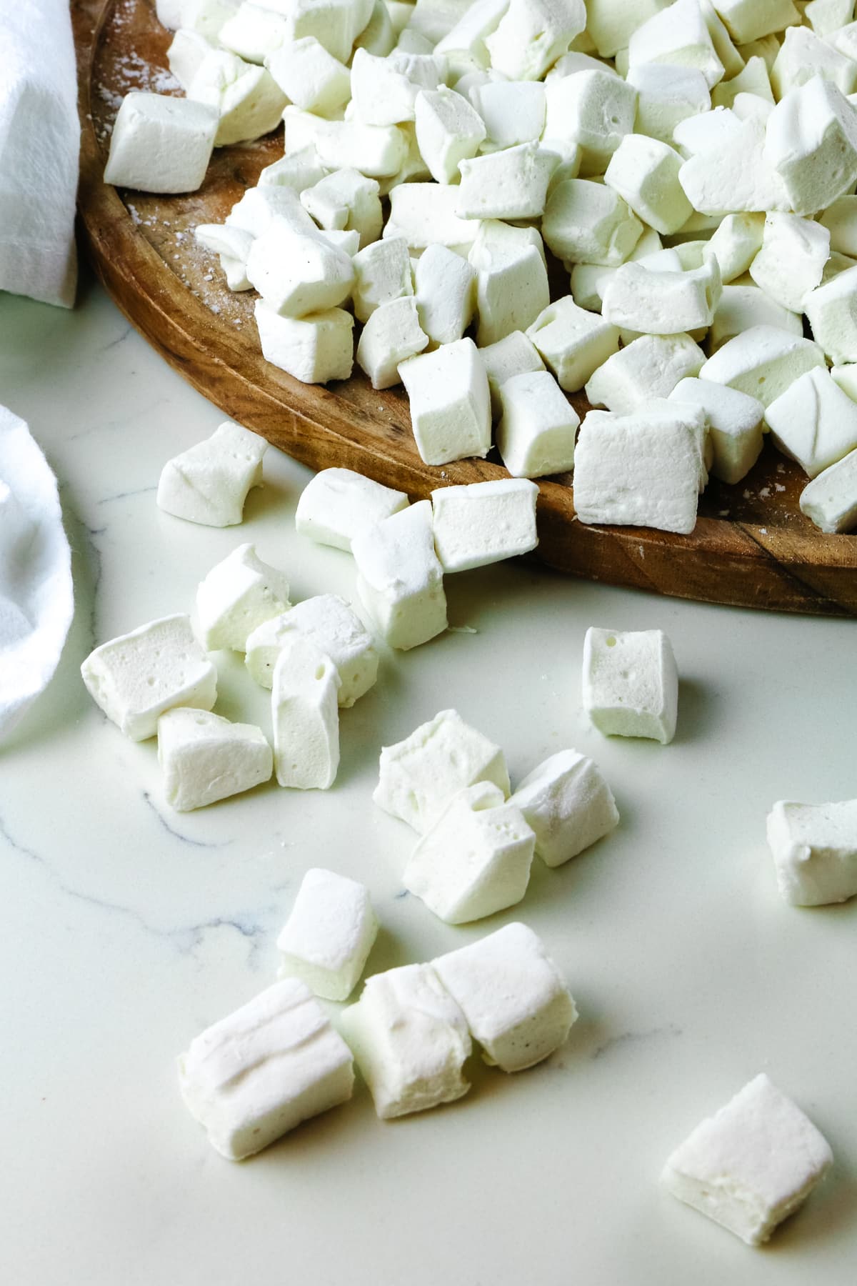 bite-sized homemade marshmallows scattered on a white countertop with a wooden cutting board on the side.