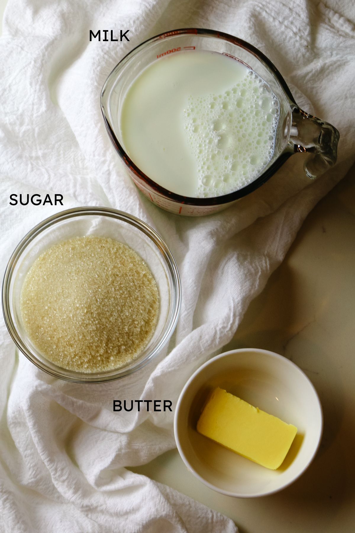 an ingredients shot including milk in a measuring cup, sugar is a glass jar, and butter in a bowl..