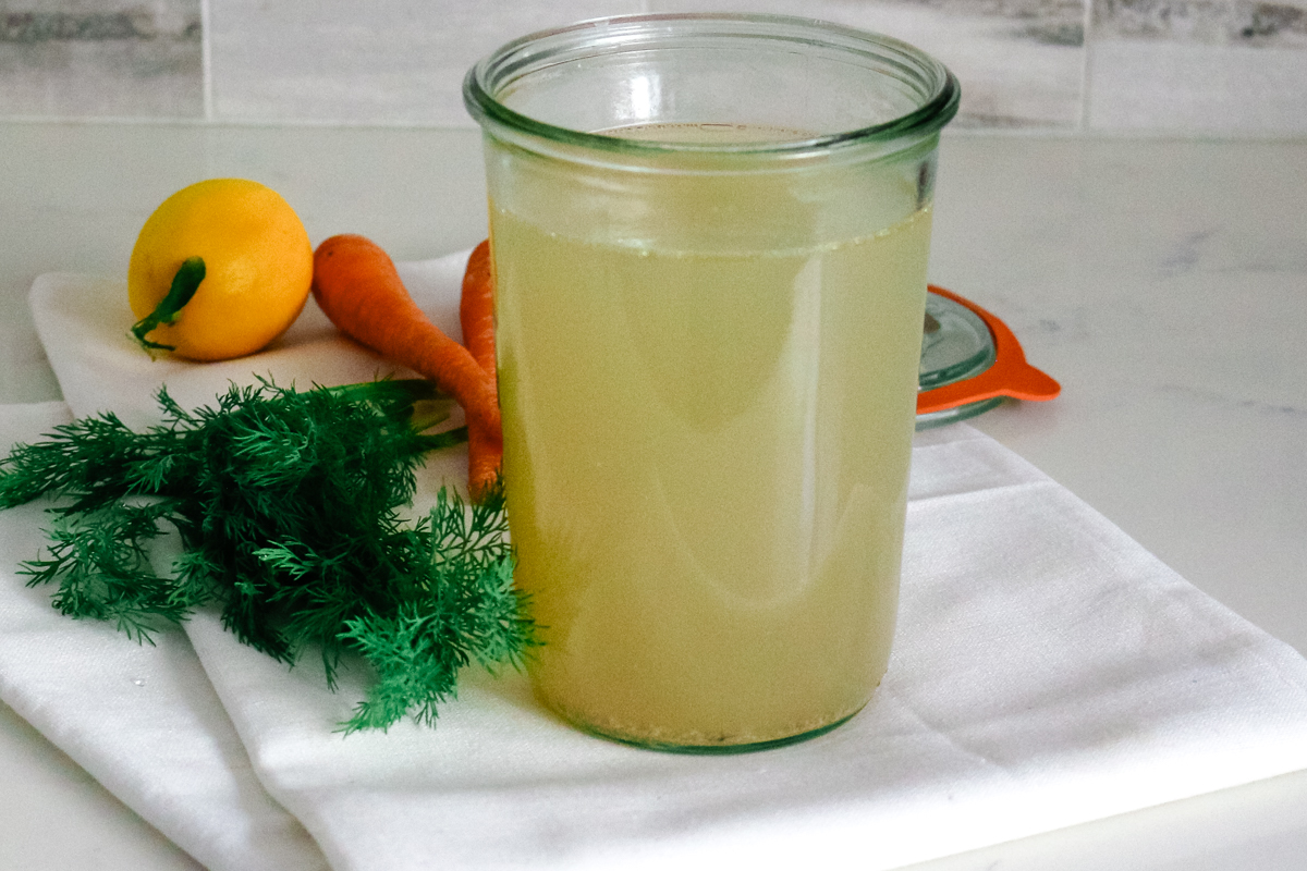 fish stock in glass jar sitting on a towel with dill, lemons, and carrots on the left of it