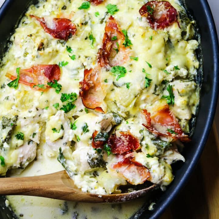 spinach artichoke and chicken in a black baking dish with a wooden spoon.