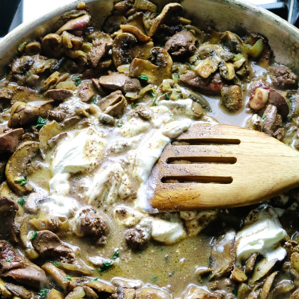 sour cream added to mushrooms and liver in a skillet with wooden spatula.