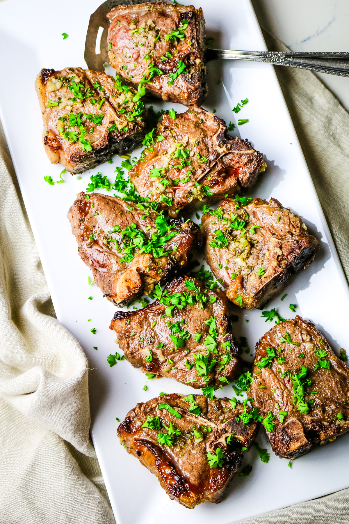 cooked lamb loin chops on a white platter with a slotted spoon and garnished with parsley.