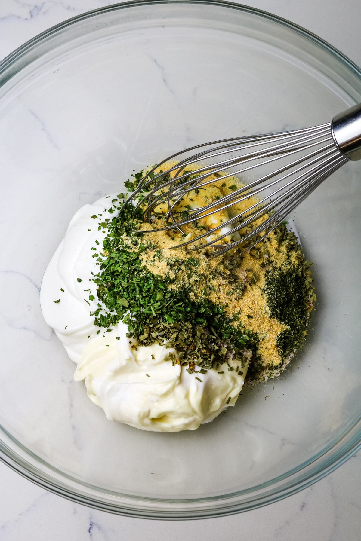 mayonnaise and Greek yogurt and dried herbs and spices in a glass mixing bowl with a whisk.