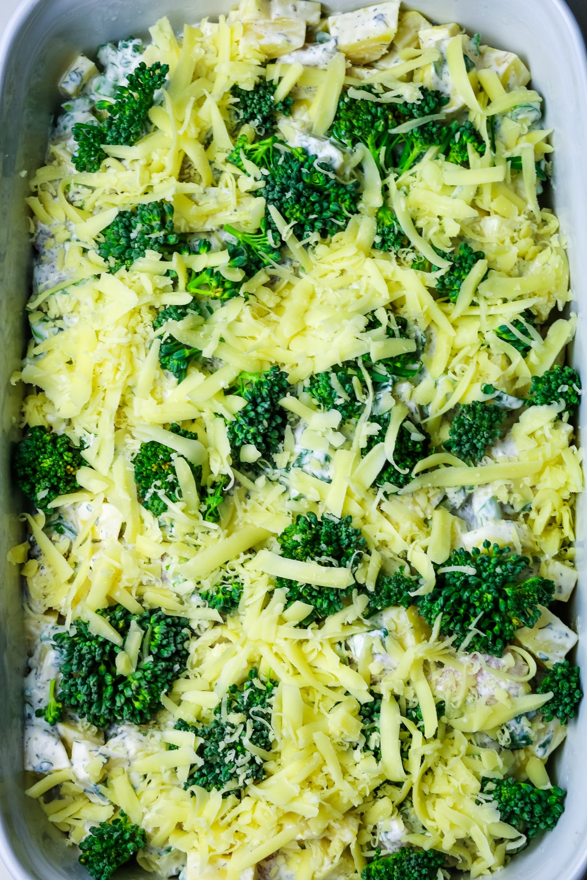 freshly grated cheese and raw broccoli florets on top of a casserole in a white baking dish.