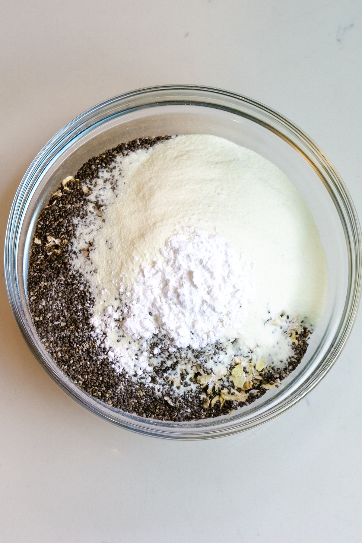 dried ingredients in a glass bowl including baking powder, oats, chia seeds, and collagen powder.