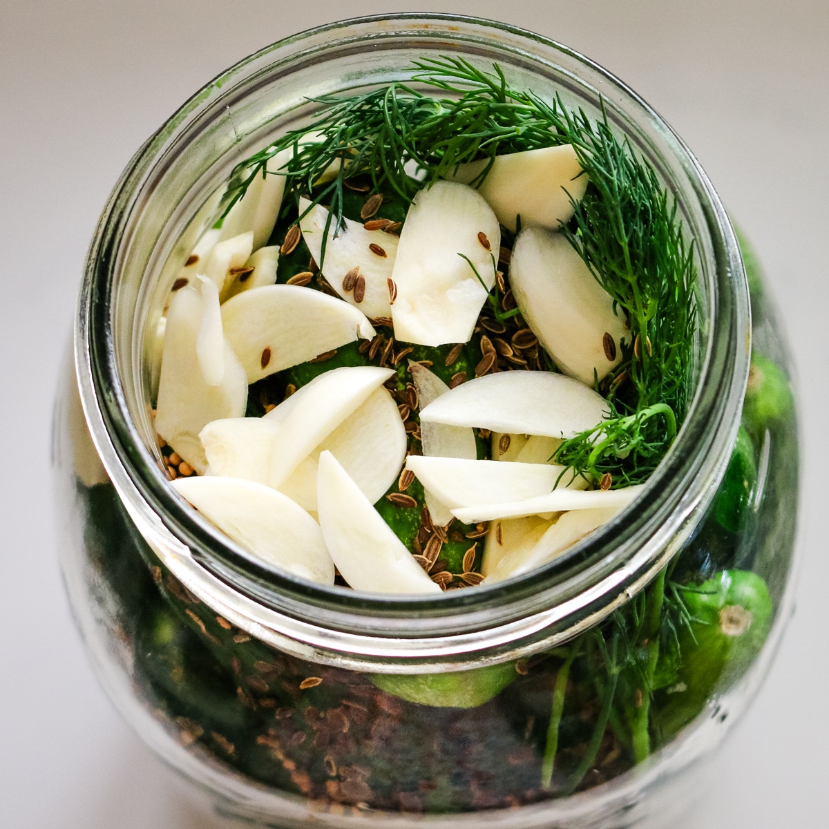 Sliced garlic and dill seeds on top of cucumbers in glass jar.