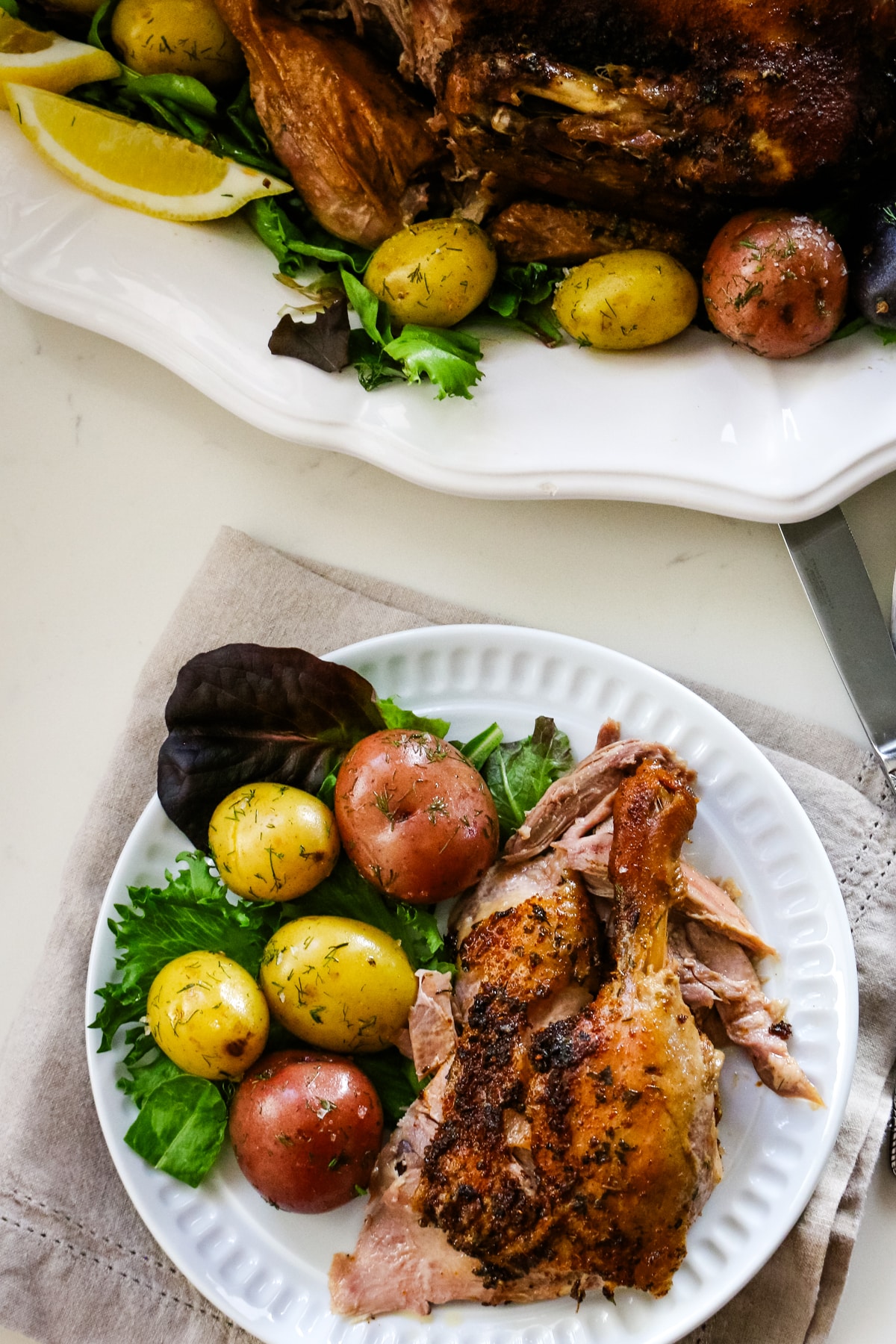 On top part of the table, it is a whole duck seasoned with garlic and marble potatoes. On another plate is a quarter-leg part of a duck on a white plate with marble potatoes and herbs.