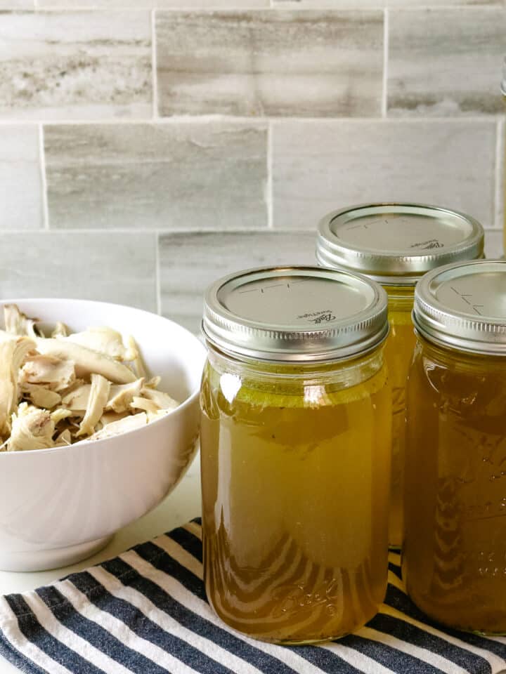 Shredded chicken in a white bowl with five jars of chicken broth beside it.
