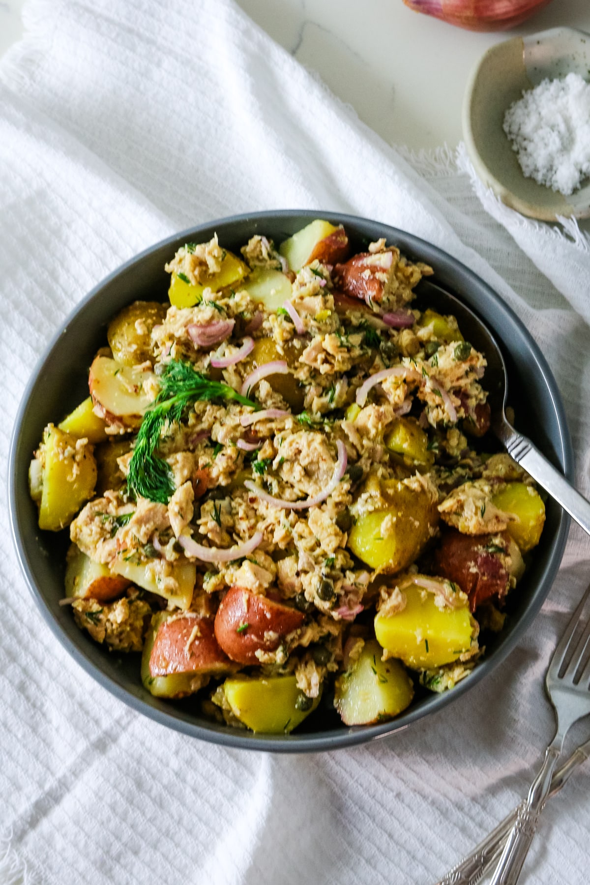 A gray bowl filled with tuna potato salad with a spoon inside and salt on the side.