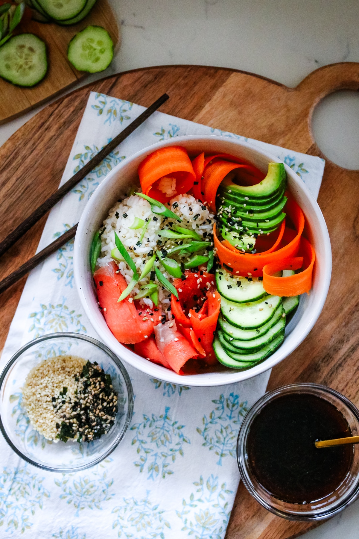 Smoked salmon in a white bowl, served with sliced cucumber, sesame seeds, and soy sauce on the side.