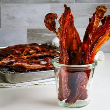 Crispy bacon strips in a clear glass jar, with more bacon cooling on a rack over a metal tray in the background.